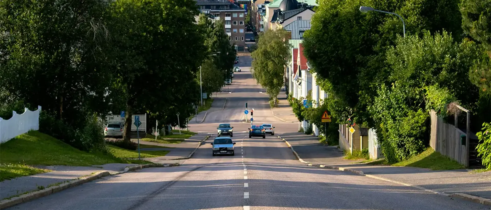 Väg i bostadsområde med bilar, staket, infrastruktur och grönska. Foto.