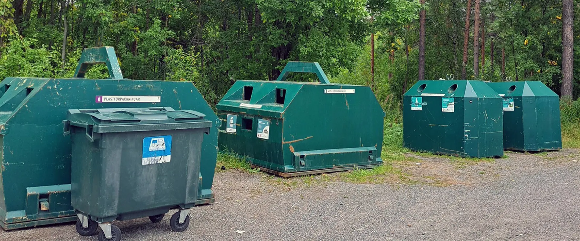 Återvinningsstation med en separat plasttunna för tidningar framför. Bilden är tagen på sommaren och träden är gröna. Foto.
