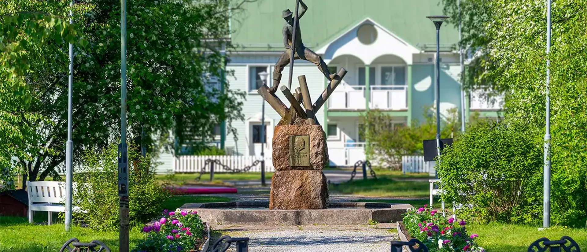 Skulpuren av en timmerflottare vid fontänen i parken i jokkmokk.  Bilden är tagen en sommardag med blommor, grönska och blå himmel. Foto.